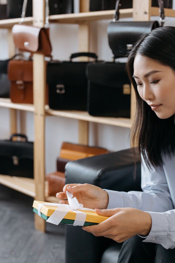 Asian woman untying a gift box in a leather bag store, showcasing various products.